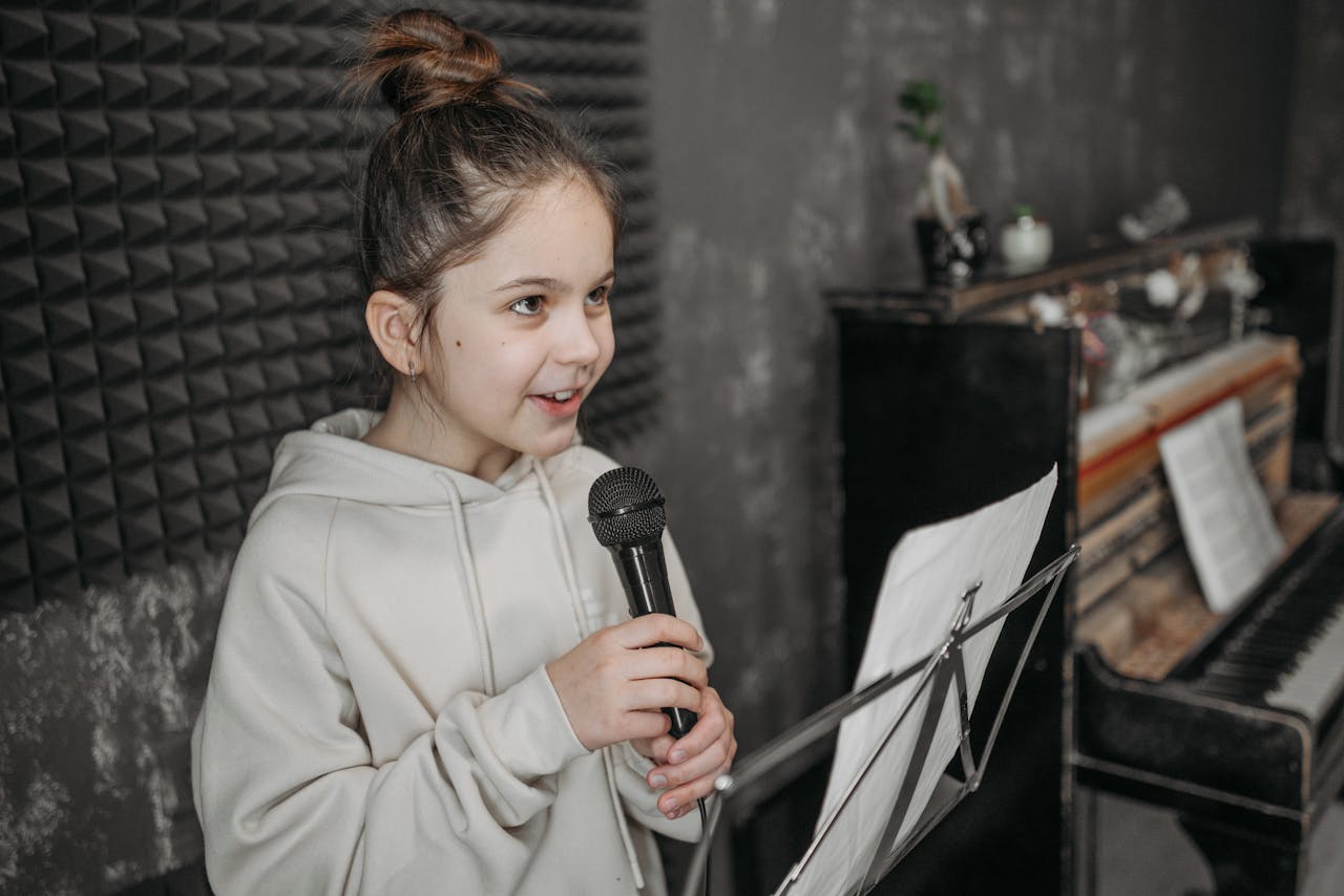 Caucasian girl singing into a microphone in a soundproof studio with sheet music and piano.